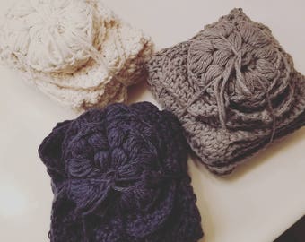May include: Three crocheted flower-shaped squares in different colors: white, gray, and dark blue. The squares are folded and stacked on top of each other.