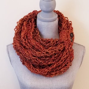 May include: A rust-colored infinity scarf with a button closure. The scarf is made of a soft, textured yarn and is displayed on a gray mannequin.