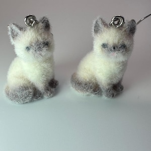 May include: A pair of cat-shaped earrings. Each earring features a small, fluffy cat figurine with a cream-colored body and gray accents on the ears, paws, and tail. The cats have small, dark eyes and pink noses. The earrings have silver-colored ear wires.