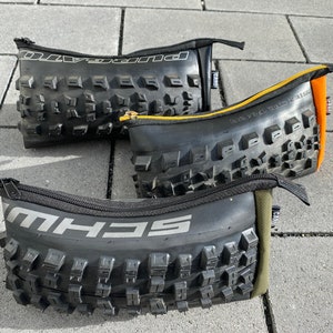 Könnte beinhalten: Drei schwarze Federmäppchen aus recycelten Fahrradreifen. Jedes Etui hat einen Reißverschluss und ist mit unterschiedlich farbigem Stoff gefüttert. Die Reifenprofile sind sichtbar, und die Markennamen sind aufgedruckt.