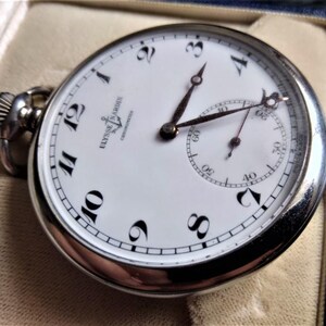 May include: A silver pocket watch with a white face, black numerals, and black hands. The watch has a subdial at the bottom and the brand name "Ulysse Nardin" is visible. The watch is in a blue box.