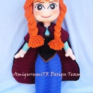 May include: A crocheted doll with red hair in braids, wearing a blue dress, a black vest, and a maroon cape. The doll has a smiling face with green eyes and is holding its hands in front of it. The doll is standing on a blue surface. The text "AmigurumiTR Design Team" is visible on the cape.