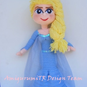 May include: A crocheted doll with long blonde hair, wearing a light blue dress with a sparkly bodice and a sheer blue cape. The doll has a smiling face with blue eyes and red lips. The doll is standing on a light blue background. The text "HAVVANIN GURUMLILERI" is at the top of the image and the text "AmigurumiTR Design Team" is at the bottom of the image.