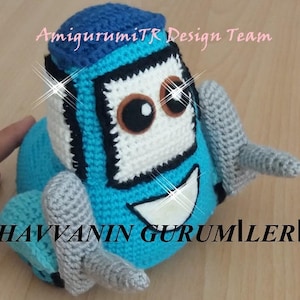 Puede incluir: Un coche de juguete de crochet azul y gris con una cara blanca y ojos marrones. El juguete está sentado sobre una superficie de madera. El texto "AmigurumiTR Design Team" es visible en el fondo.