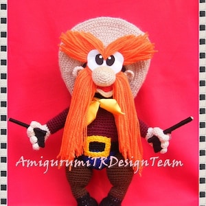 May include: A crocheted plush toy of a cartoon character with a red shirt, yellow belt, and orange hair. The character is holding two toy guns and has a brown hat. The text "havvannin gurumileri" and "Amigurumi TR Design Team" are visible in the image.