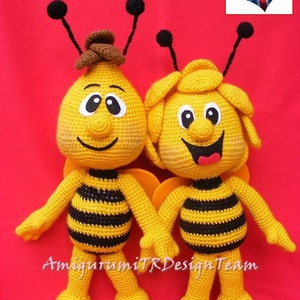 May include: Two crocheted bee plushes, one with a brown hat, standing side-by-side on a red background. The bees are yellow with black stripes and have black eyes and smiling faces. The text "AmigurumiTRDesignTeam" is visible at the bottom of the image.