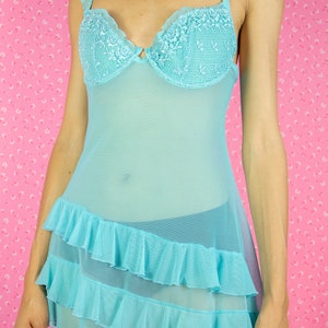 The Y2K Blue Mesh Slip Dress is a vintage pastel blue semi-sheer babydoll featuring ruffles, adjustable straps adorned with rhinestones, floral details, and a cut-out back with a bow. Romantic coquette 2000s lingerie cami night dress.