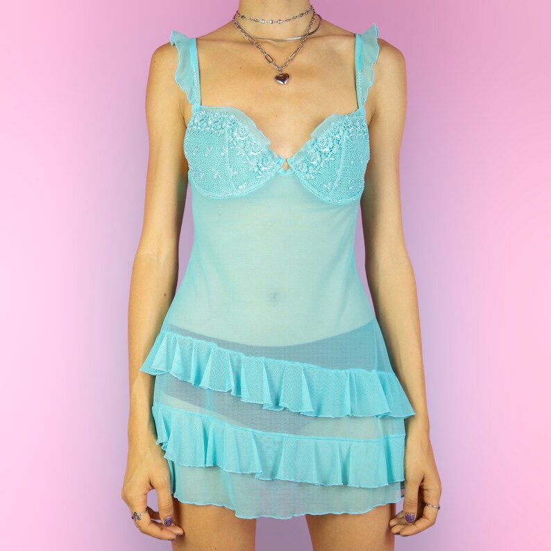 The Y2K Blue Mesh Slip Dress is a vintage pastel blue semi-sheer babydoll featuring ruffles, adjustable straps adorned with rhinestones, floral details, and a cut-out back with a bow. Romantic coquette 2000s lingerie cami night dress.