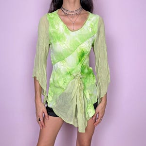 May include: A lime green long-sleeve top featuring a V-neckline, sheer fabric with a crinkled texture, and a subtle floral pattern. The garment includes a decorative fabric tie detail at the front hem, creating an asymmetrical drape.