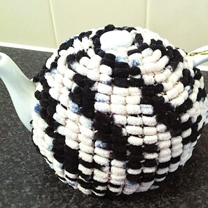 May include: A white ceramic teapot covered by a thick, textured crochet cozy. The cozy features a repeating pattern of black and white yarn loops, with subtle hints of blue throughout the material. The teapot has a curved handle and spout.