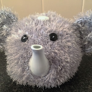 May include: A novelty teapot in the shape of an elephant's head. The teapot is covered in soft, grey, fluffy yarn, with large black button eyes and light blue yarn ears. The spout and handle are white ceramic.