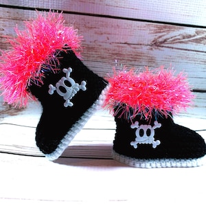 May include: A pair of black crochet baby boots with a white skull and crossbones design. The boots have a fluffy pink trim and a white base. The boots are displayed on a light wood background.
