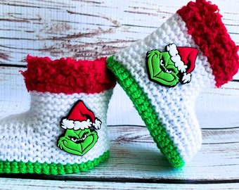 May include: A pair of white crocheted booties featuring a bright green sole and a textured red cuff. Each bootie is decorated with a small, colorful rubber emblem of a green character wearing a red Santa hat, positioned on the side of the white knit.