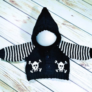 May include: A black hooded cardigan featuring long sleeves with white horizontal stripes. The front of the garment displays two white skull and crossbones motifs, one on each side of the central button closure, knitted into the black fabric.