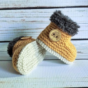Peut inclure: Une paire de chaussons de bébé crochetés à la main. Les chaussons sont jaunes avec une bordure en fausse fourrure grise et un bouton en bois avec un motif d'arbre. Les semelles sont de couleur crème.
