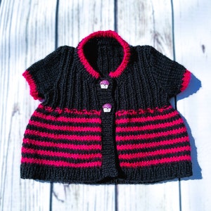 May include: A black and pink striped knitted baby cardigan with short sleeves and two cupcake buttons.