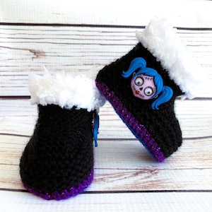 May include: A pair of black crocheted baby boots with white faux fur trim. The boots have a purple and blue trim and a decorative face with blue pigtails. The boots are displayed on a light wood surface.