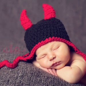 May include: A sleeping baby wearing a handmade crochet devil hat. The hat is black with red trim and horns. The baby is resting on a grey surface.