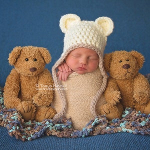 May include: A newborn baby wearing a white crocheted hat with bear ears, swaddled in a beige blanket, is surrounded by two brown teddy bears on a blue and brown fuzzy blanket.