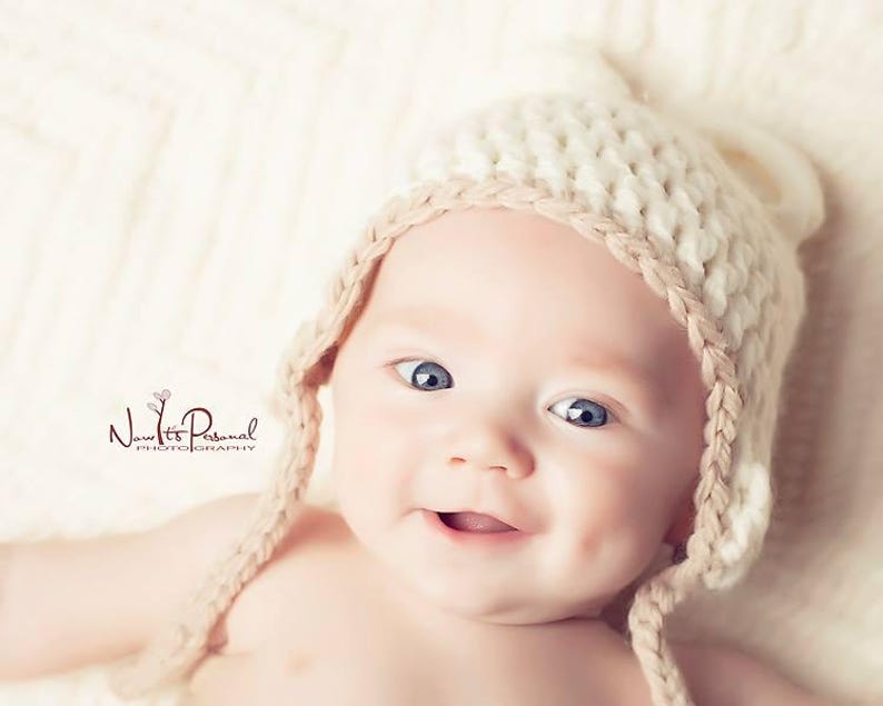 K&ouml;nnte beinhalten: Ein Baby tr&auml;gt eine cremefarbene, grob gestrickte M&uuml;tze mit Ohrenklappen und einem hellbraunen geflochtenen Rand. Die M&uuml;tze hat kleine, abgerundete Ohren an der Oberseite. Das Baby hat leuchtend blaue Augen und ein sichtbares Gr&uuml;bchen an der Wange.
