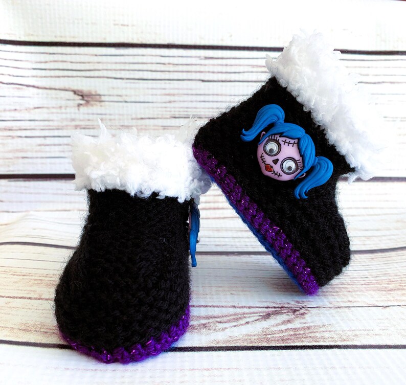 May include: A pair of black crocheted baby boots with white faux fur trim. The boots have a purple and blue trim and a decorative face with blue pigtails. The boots are displayed on a light wood surface.