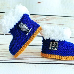 May include: A pair of handmade baby booties. The booties are blue with white faux fur trim and a gold-colored sole. One bootie has an owl pin, and the other has a pin that says "Book of Spells".