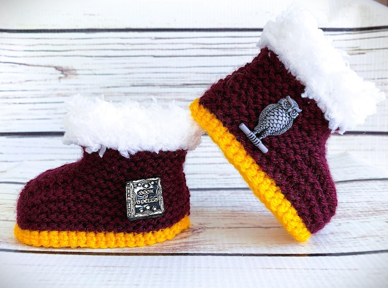 May include: A pair of maroon crocheted baby booties with yellow trim and white fur. One bootie has a silver owl charm and the other has a silver charm with the words "Book of Spells".