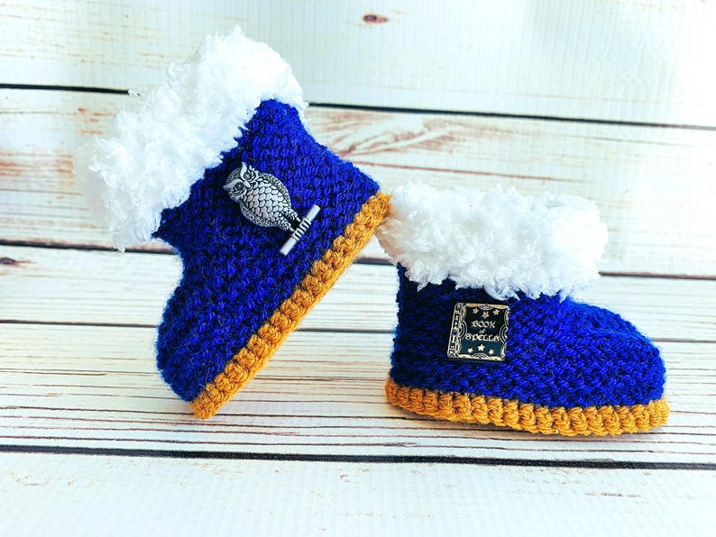 May include: A pair of handmade baby booties. The booties are blue with white faux fur trim and a gold-colored sole. One bootie has an owl pin, and the other has a pin that says "Book of Spells".