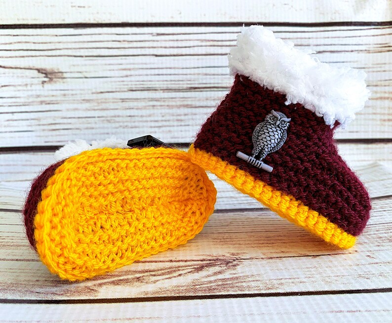 May include: Crocheted baby booties and matching taco-shaped item. The booties are burgundy with a yellow trim and white faux fur trim. A silver owl pin adorns the side. The taco-shaped item is yellow with burgundy trim.