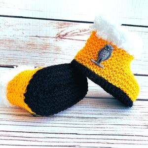 May include: A pair of handmade crochet baby booties. The booties are yellow with black soles and white faux fur trim. Each bootie has a silver owl embellishment. The booties are displayed on a light wood surface.