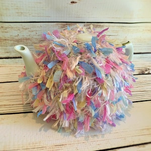 May include: A white ceramic teapot covered in a colorful, fluffy fabric tea cozy. The cozy is made of pink, blue, yellow, and cream-colored fabric strips. The teapot has a spout, handle, and lid.