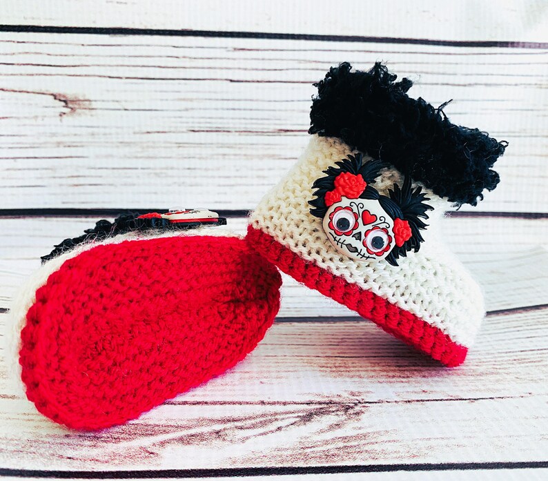 Puede incluir: Un par de botines de beb&eacute; de ganchillo hechos a mano. Los botines son blancos con ribetes rojos y presentan un dise&ntilde;o de calavera del D&iacute;a de Muertos. La parte superior de los botines tiene ribetes negros. La suela de los botines es roja.