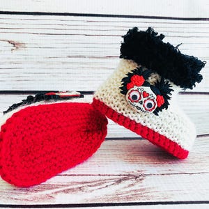Puede incluir: Un par de botines de beb&eacute; de ganchillo hechos a mano. Los botines son blancos con ribetes y suelas rojas. Cada bot&iacute;n presenta una aplicaci&oacute;n de calavera del D&iacute;a de Muertos con rosas rojas y pelo negro. La parte superior del bot&iacute;n tiene un ribete negro.