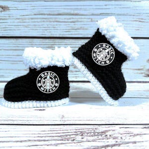 May include: A pair of black and white crocheted baby boots. Each boot features a white trim and a white patch with the words "BASIC WITCH" and a witch logo. The boots are displayed on a light blue and white wooden surface.