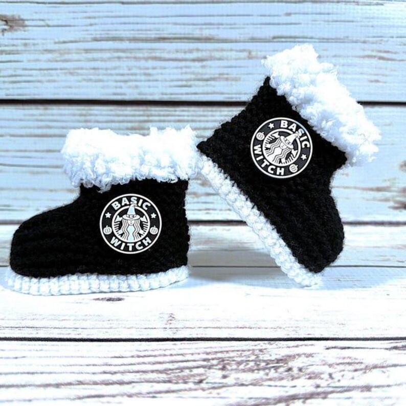 May include: A pair of black and white crocheted baby boots. Each boot features a white trim and a white patch with the words "BASIC WITCH" and a witch logo. The boots are displayed on a light blue and white wooden surface.