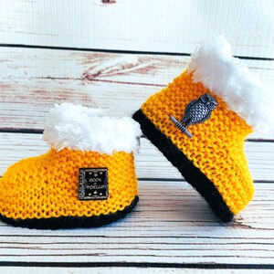 May include: A pair of handmade yellow knitted baby booties with black soles and white faux fur trim. One bootie has a small metal owl charm, and the other has a small square patch with the words "Book of Spells".
