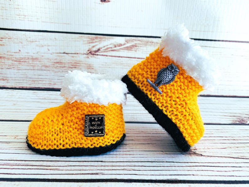 May include: A pair of handmade yellow knitted baby booties with black soles and white faux fur trim. One bootie has a small metal owl charm, and the other has a small square patch with the words "Book of Spells".