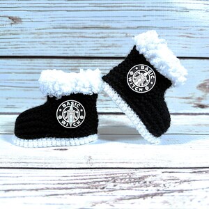 May include: A pair of black and white crocheted baby boots. The boots have a white fluffy trim and a white stitched sole. Each boot features a white circular logo with the words "Basic Witch".