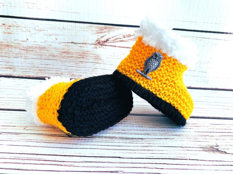 May include: A pair of handmade crochet baby booties. The booties are yellow with black soles and white faux fur trim. Each bootie has a silver owl embellishment. The booties are displayed on a light wood surface.