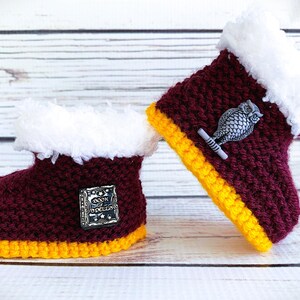 May include: A pair of burgundy and yellow crocheted baby boots with white faux fur trim. One boot features a silver owl pin, the other a silver pin with the text "Book of Spells".