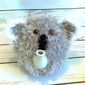 May include: A koala-shaped tea cozy made of fuzzy gray and white yarn. The item features two large, rounded ears, dark brown plastic eyes, and a prominent black nose. A white ceramic teapot spout protrudes from the center of the fuzzy face.