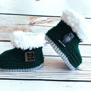 May include: A pair of handmade, dark green crochet baby booties with white faux fur trim. One bootie has a small metal owl charm, the other has a metal tag with the text "Book of Spells". The booties have a gray trim and are on a wooden surface.