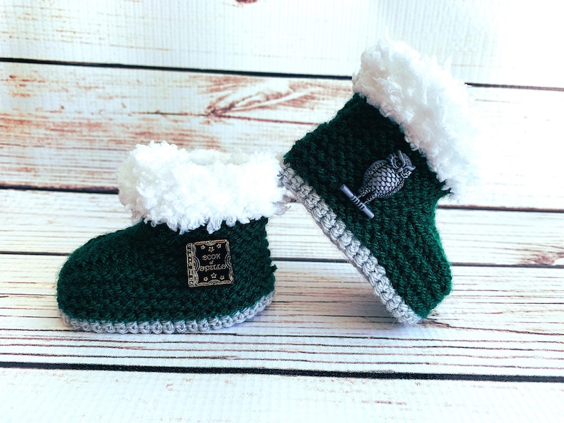 May include: A pair of handmade, dark green crochet baby booties with white faux fur trim. One bootie has a small metal owl charm, the other has a metal tag with the text "Book of Spells". The booties have a gray trim and are on a wooden surface.