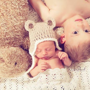 K&ouml;nnte beinhalten: Eine beige geh&auml;kelte B&auml;renm&uuml;tze mit abgerundeten Ohren und wei&szlig;em Wellensaum, getragen von einem Neugeborenen, das neben einem gro&szlig;en, flauschigen, hellbraunen Teddyb&auml;ren und einem kleinen Kind auf einer strukturierten, cremefarbenen Decke liegt.