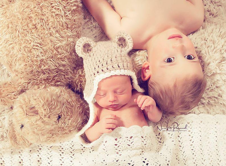 K&ouml;nnte beinhalten: Eine beige geh&auml;kelte B&auml;renm&uuml;tze mit abgerundeten Ohren und wei&szlig;em Wellensaum, getragen von einem Neugeborenen, das neben einem gro&szlig;en, flauschigen, hellbraunen Teddyb&auml;ren und einem kleinen Kind auf einer strukturierten, cremefarbenen Decke liegt.
