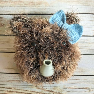 May include: A brown fuzzy teapot with a light blue crocheted bow on top. The teapot has a white ceramic spout.