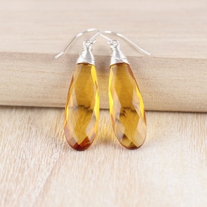 May include: A pair of drop earrings featuring faceted, teardrop-shaped amber-colored stones. Each stone is secured with silver-toned wire wrapping at the top, which connects to a simple silver-toned hook ear wire for hanging the jewelry piece.