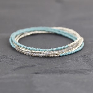 May include: A multi-strand bracelet featuring a combination of small, matte light blue beads, metallic silver-toned beads, and faceted, translucent grey gemstone beads arranged in a layered, circular design with a delicate, thin profile.