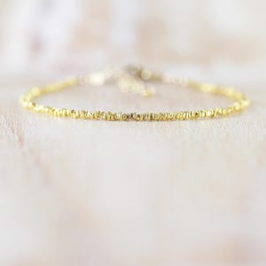 Peut inclure: Un bracelet chaîne doré délicat composé d'une série de petites perles métalliques texturées et irrégulières enfilées en une ligne continue, créant un effet scintillant subtil sur toute la longueur de ce bijou fin.