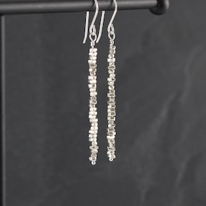 Könnte beinhalten: Ein Paar lange, hängende Silberohrringe mit einem vertikalen Strang aus kleinen, unregelmäßigen, metallischen, nuggetförmigen Perlen. Jeder Ohrring hängt an einem einfachen Silberhaken und erzeugt eine schlanke, lineare Silhouette mit texturierter Oberfläche.